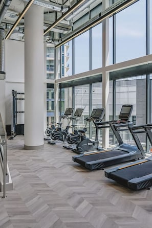 Een fitnessruimte met loopbanden en hometrainers, grote ramen en een visgraatvloer.
