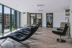 A modern waiting area with a row of black chairs, a visible exit sign, and a view of the outside buildings through large windows. - The Aqua Vista | 2 Bed, Lake Views w Pool, Hot tub (Kelowna)