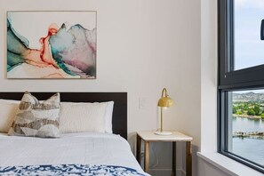 A bedroom with a bed, pillows, a bedside table with a lamp, and a painting on the wall. - The Aqua Vista | 2 Bed, Lake Views w Pool, Hot tub (Kelowna)