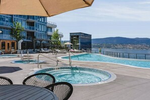 A rooftop pool area with lounge chairs, a hot tub, and a view of the city and mountains. - The Aqua Vista | 2 Bed, Lake Views w Pool, Hot tub (Kelowna)