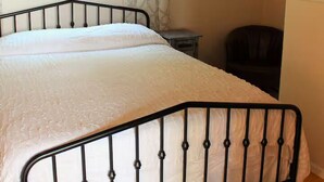 Un dormitorio con estructura metálica en la cama, una mesita de noche, una silla, una ventana con cortinas y un espejo en la pared.