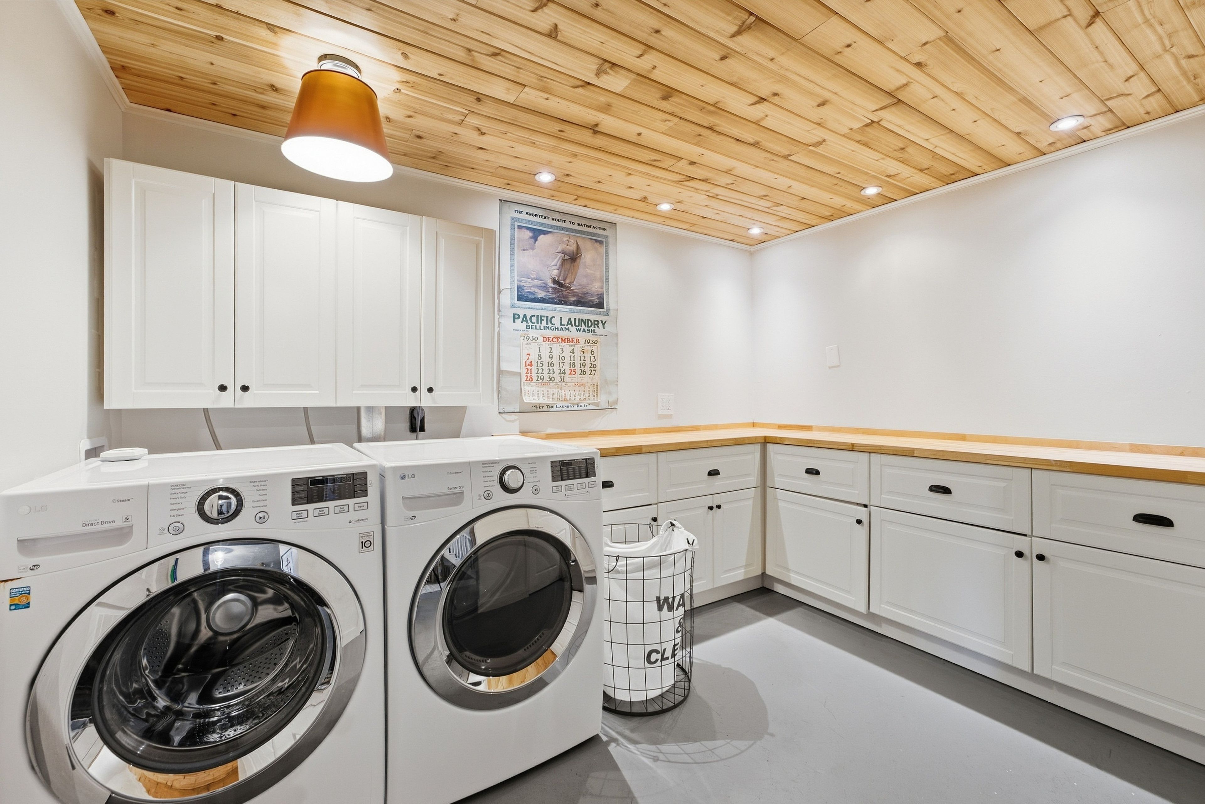 A laundry room with two front-loading washing machines, a drying rack, and a wooden ceiling.