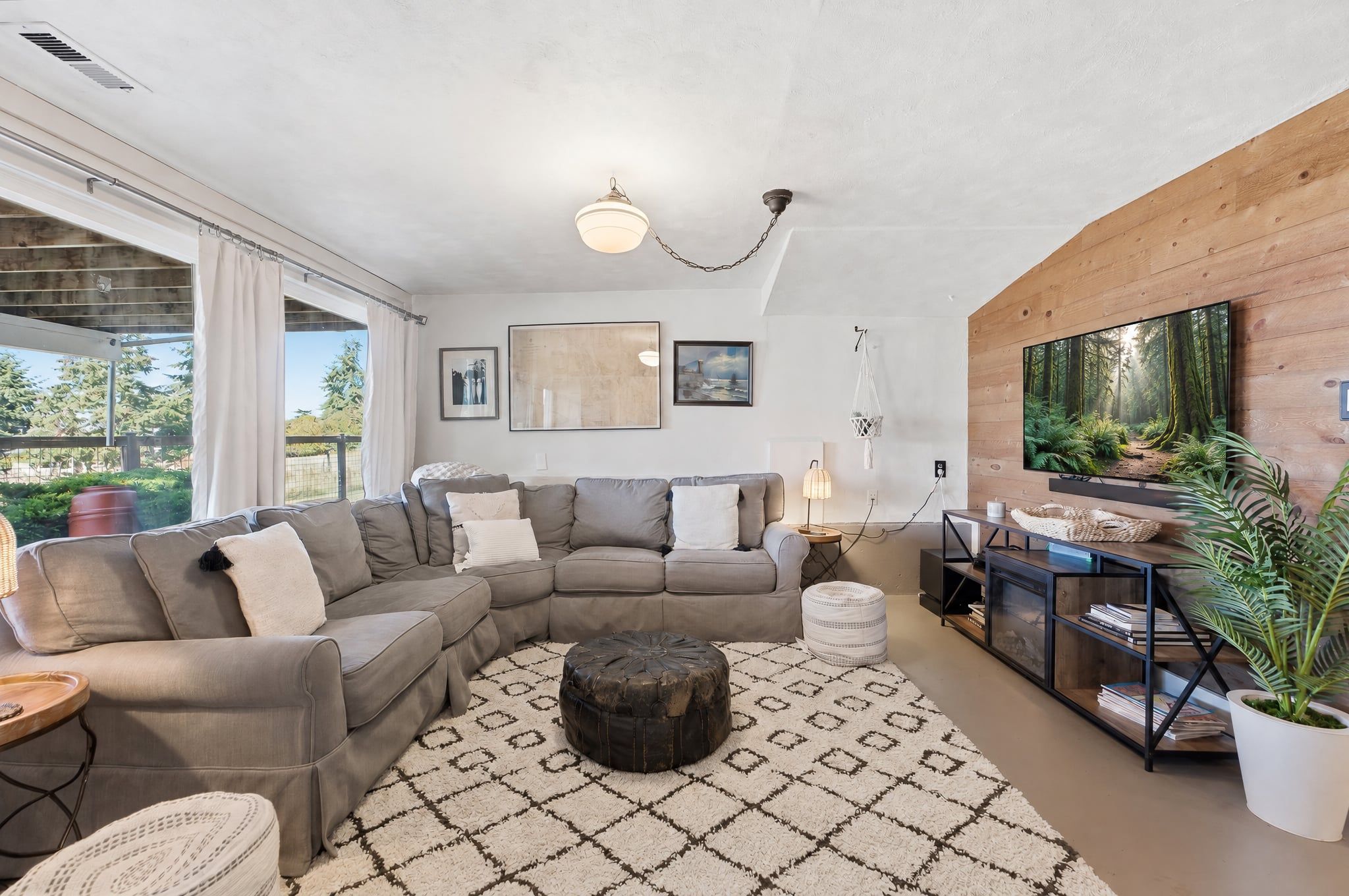 A modern living room with a sectional sofa, a round ottoman, a patterned rug, a wooden TV stand, and a large window with curtains.