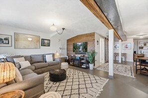 A living room with a sofa, ottoman, and a television. There is a wooden ceiling and a fireplace.