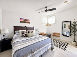Ein modernes Schlafzimmer mit einem Bett, Nachttischen, einem Schreibtisch, einem Sessel, einer Stehlampe, einem Spiegel und einer Topfpflanze.