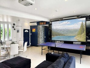 Ein modernes Wohnzimmer mit einem großen Flachbildfernseher, einem Tischtennisch und einem Essbereich mit weißem Tisch und Stühlen.