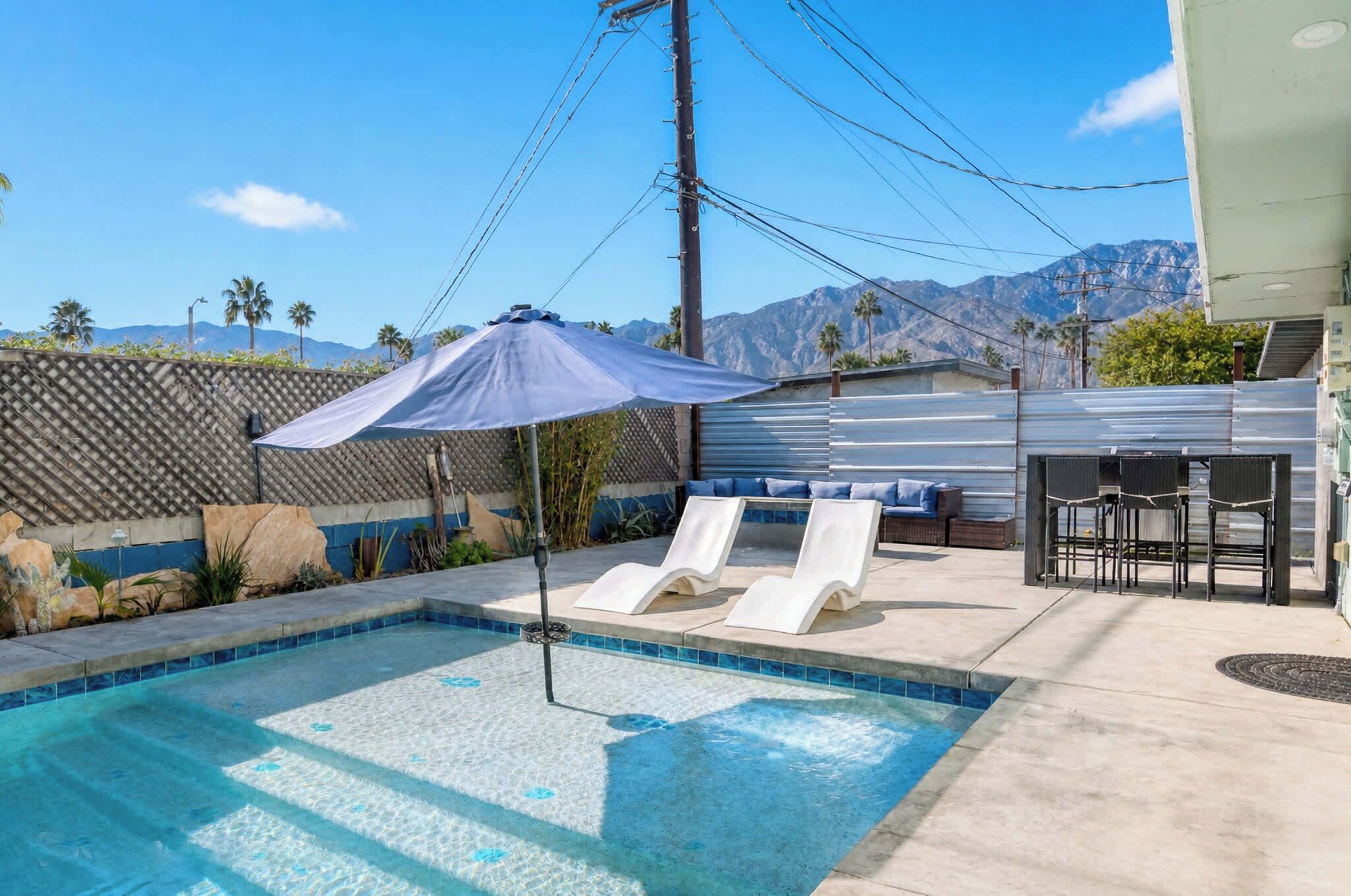 A swimming pool with lounge chairs and a patio area, surrounded by mountains and palm trees.