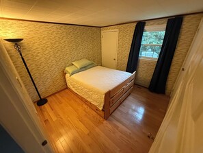 Ein Schlafzimmer mit Holzboden, einem Bett mit Steppdecke, einem Nachttisch, einer Lampe, einem Fenster mit Vorhängen und einer Tür.