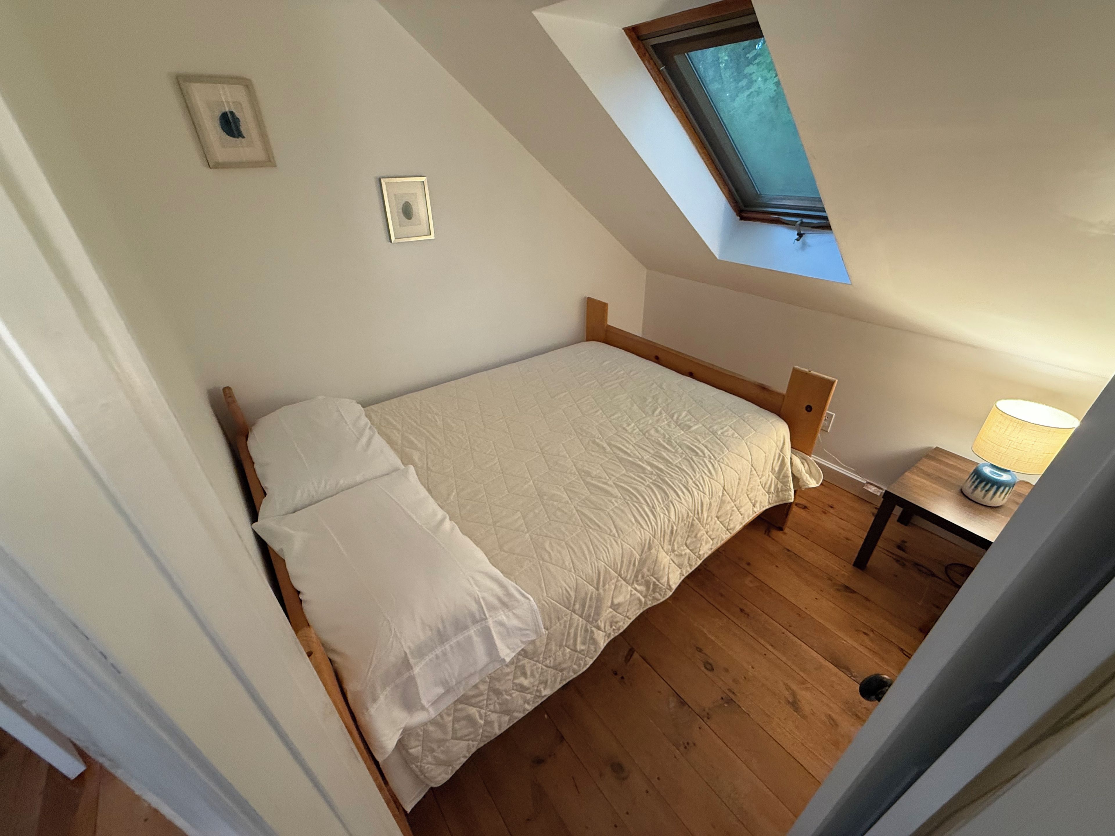A single bed with a wooden headboard in a room with wooden flooring, a small bedside table, and a window with a view of trees.