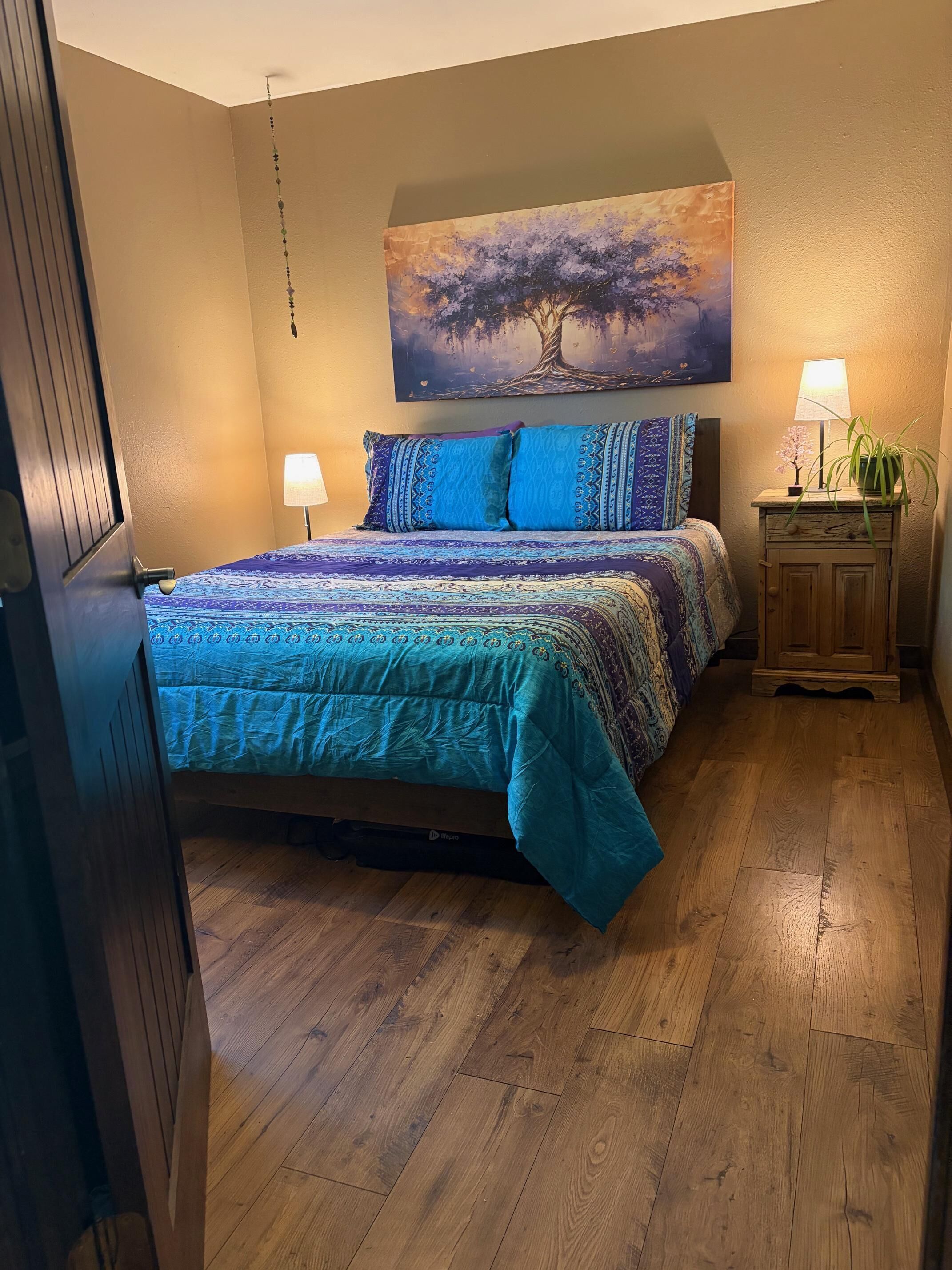 Un dormitorio con cama, mesita de noche, lámpara y un cuadro en la pared.