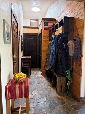 A hallway with a stone-tiled floor, wooden walls, a coat rack with jackets, a table with a basket, and a door with a handle. - 3-bedroom, 2-bath gated community of Blue River Valley Ranch Lakes (Silverthorne)