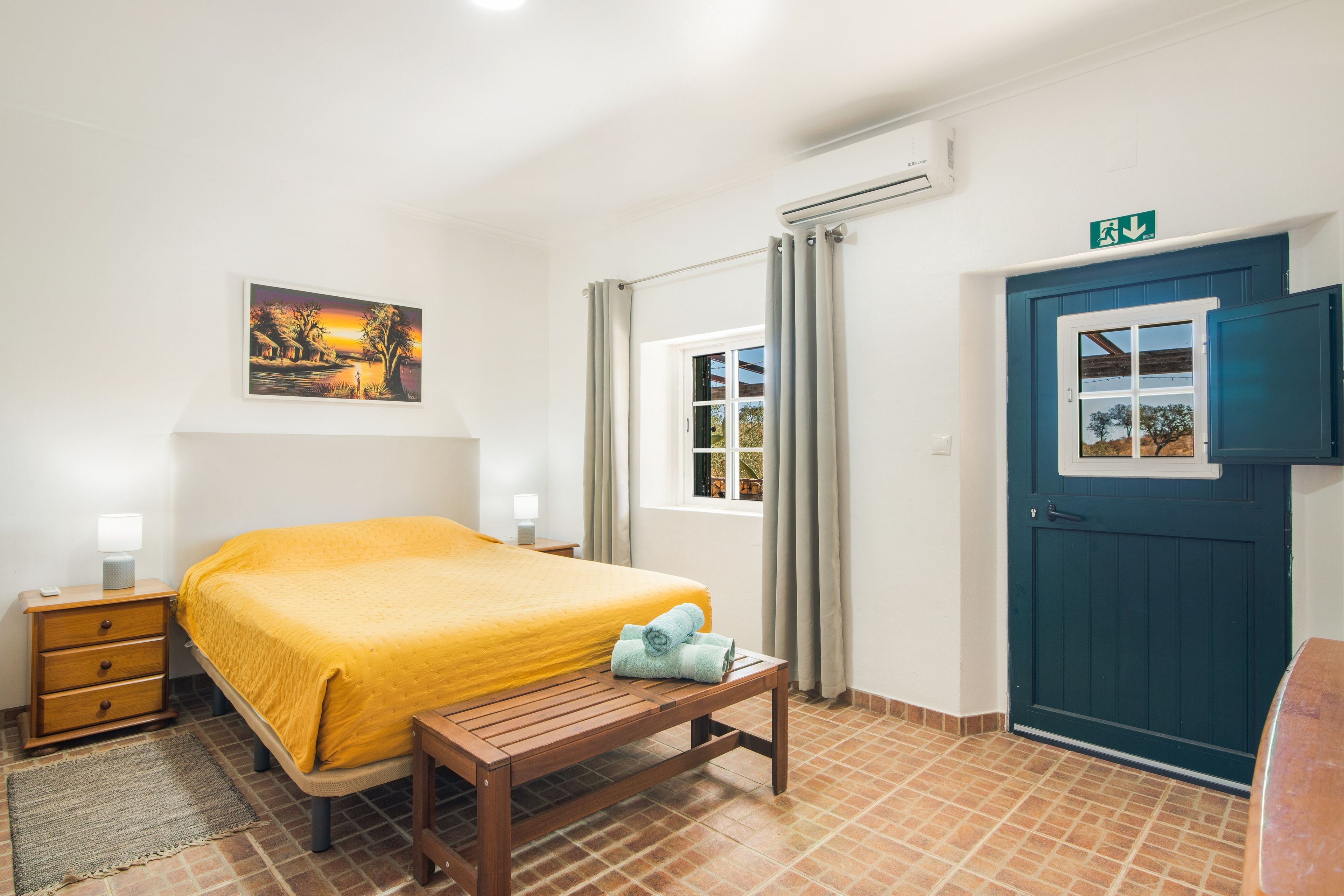 Habitación de hotel con cama, banqueta, mesita de noche, un cuadro, ventana con cortinas y puerta con vista al exterior.