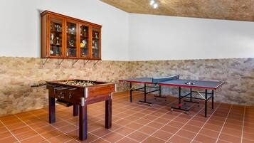Una habitación con mesa de fútbolín, mesa de ping pong y una vitrina con trofeos, todo frente a una pared de piedra.