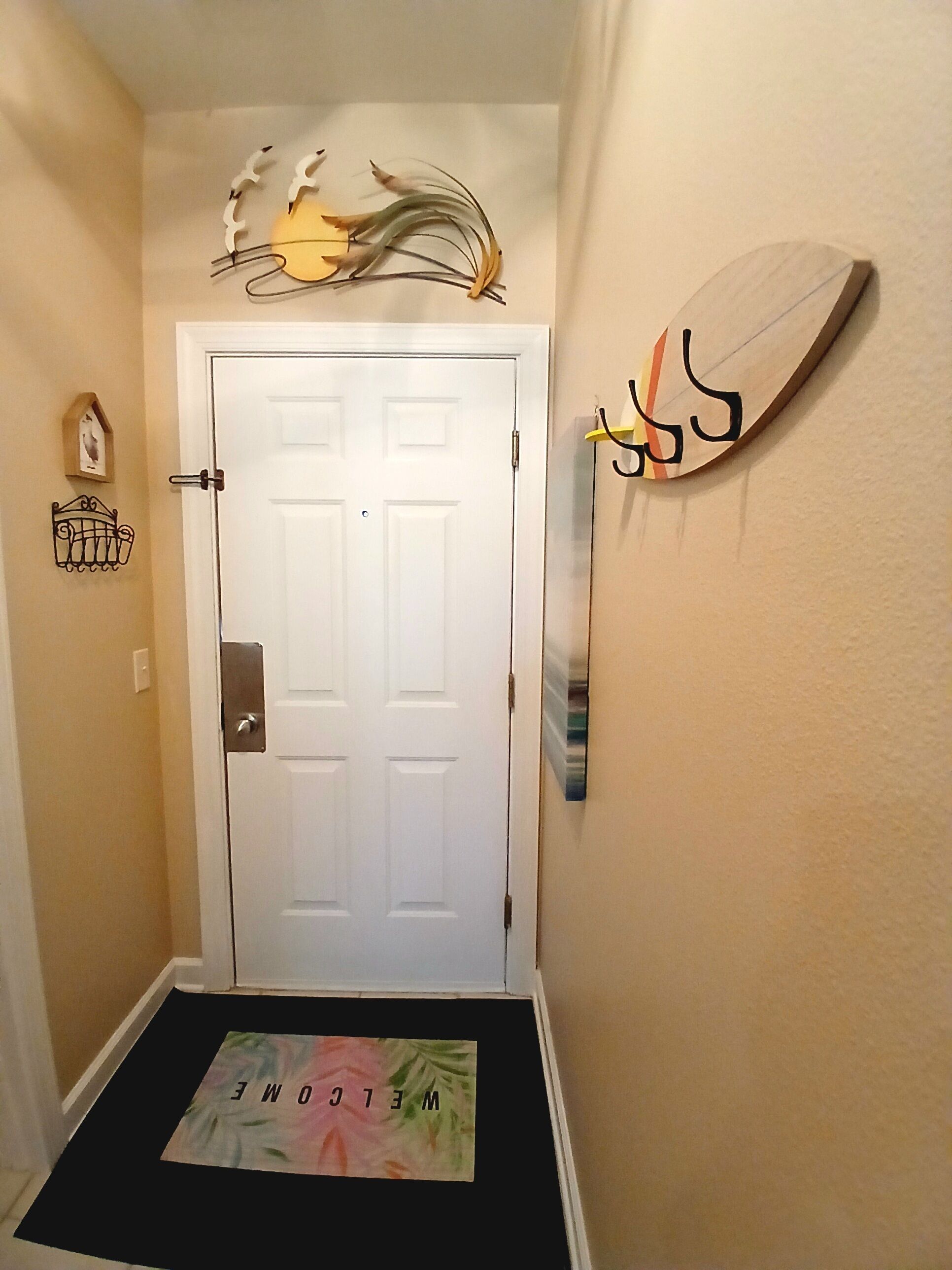A hallway with a white door, a welcome mat, and wall decorations including a birdhouse and a wall-mounted art piece.