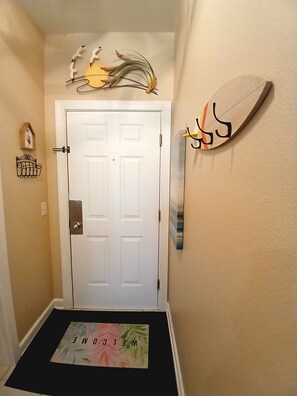 A hallway with a white door, a welcome mat, and wall decorations including a birdhouse and a wall-mounted art piece. - Sunset Beach Sea Trail Champions suite!  New and nice! Great 1st floor location (Sunset Beach)