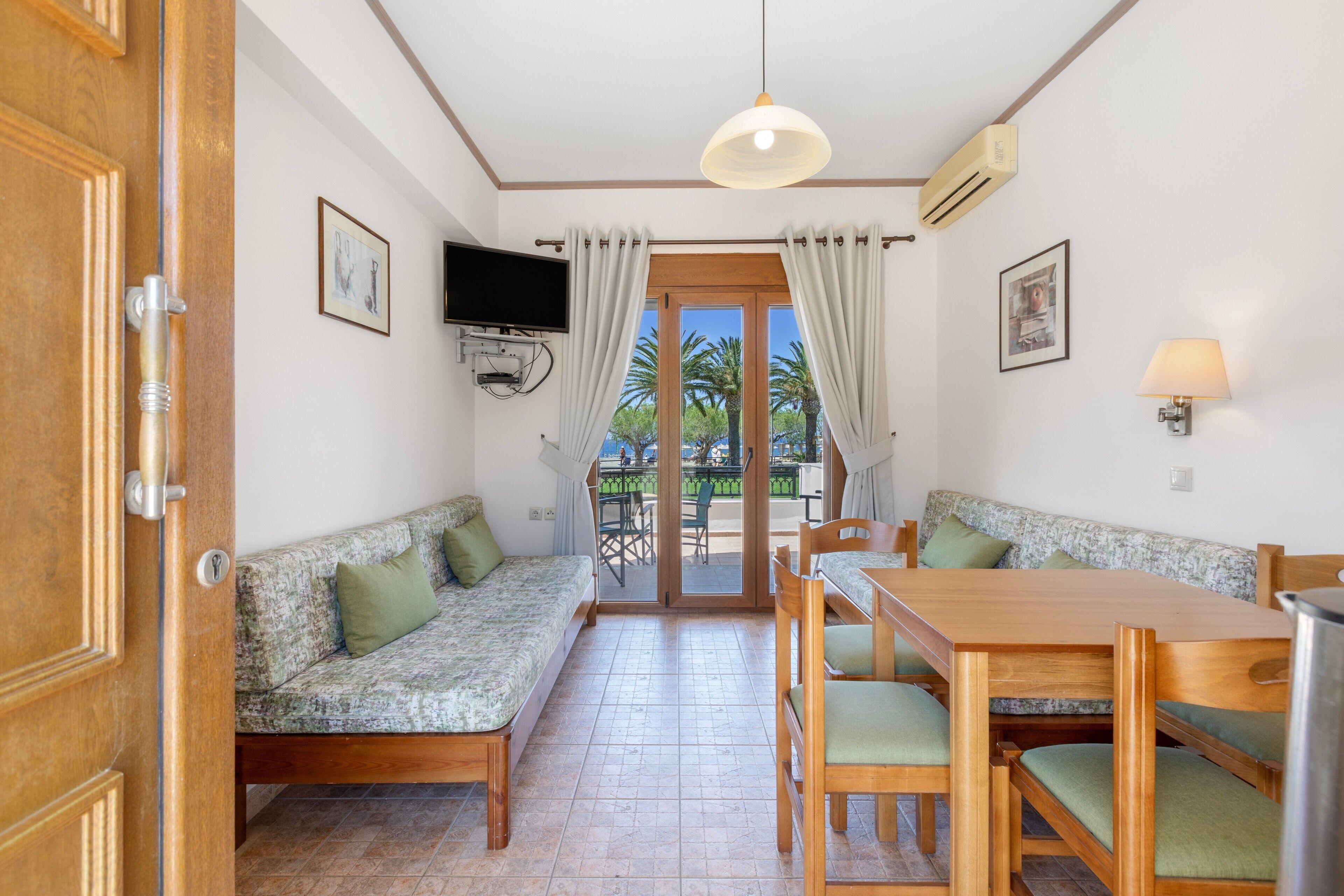 A room with a sofa, a dining table, chairs, a TV mounted on the wall, and a view of palm trees through the door.