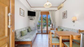 A room with a sofa, a dining table, chairs, a TV mounted on the wall, and a view of palm trees through the door.