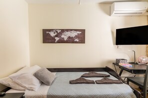 Ein Hotelzimmer mit einem Bett, Kissen, einer Weltkarte als Wandbild, einem Fernseher und einem Schreibtisch mit Ventilator und Büchern.