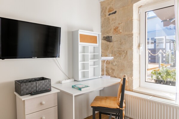 A room with a white desk, a black television mounted on the wall, a white shelving unit, a small table with a lamp, a chair, and a window with a view of the outside.
