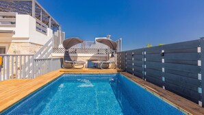Ein Schwimmbecken mit Holzterrasse, zwei Liegestühlen und Blick auf ein Gebäude mit Balkon.