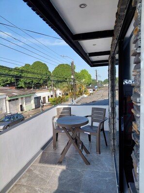 Uma varanda com mesa e cadeiras, vista para a rua e um edifício com telhado em azulejos.
