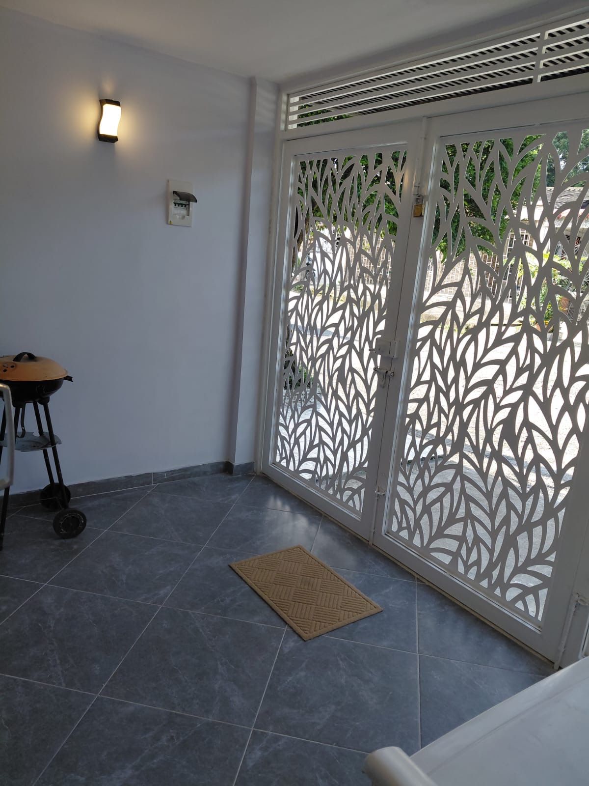 Une pièce avec un sol carrelé, une porte blanche ornée de motifs découpés, un luminaire fixé au mur et une petite table avec une chaise.