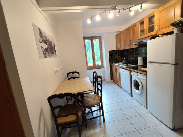 A compact kitchen with a dining table, chairs, a washing machine, and a refrigerator. - Gîtes Noryema: Bright and Functional 27m² T2, Garden by the River, Calm Setting (Entraunes)