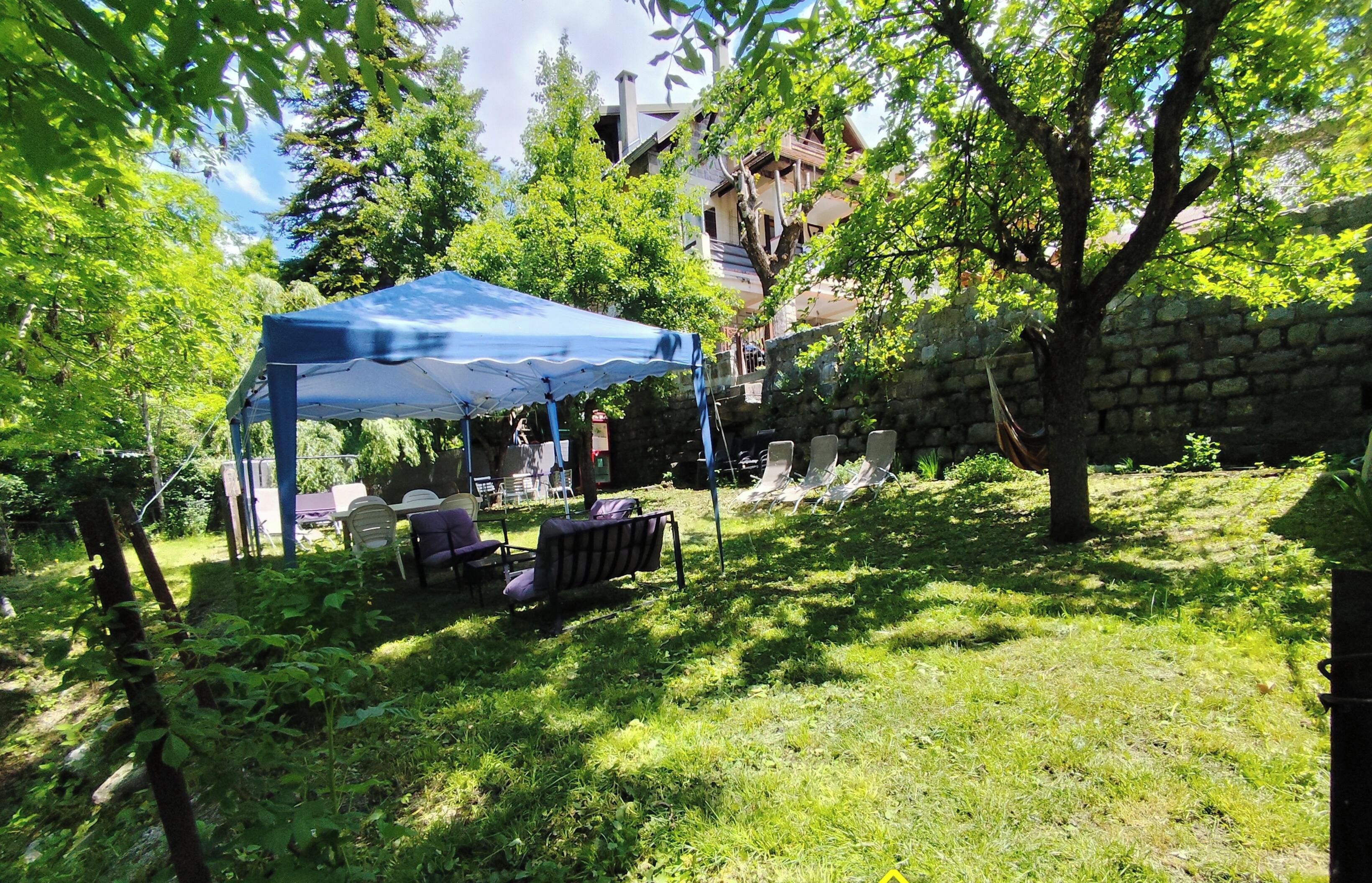 Ein überdachter Außenbereich mit Stühlen und Tisch, umgeben von Bäumen und einer Steinmauer.