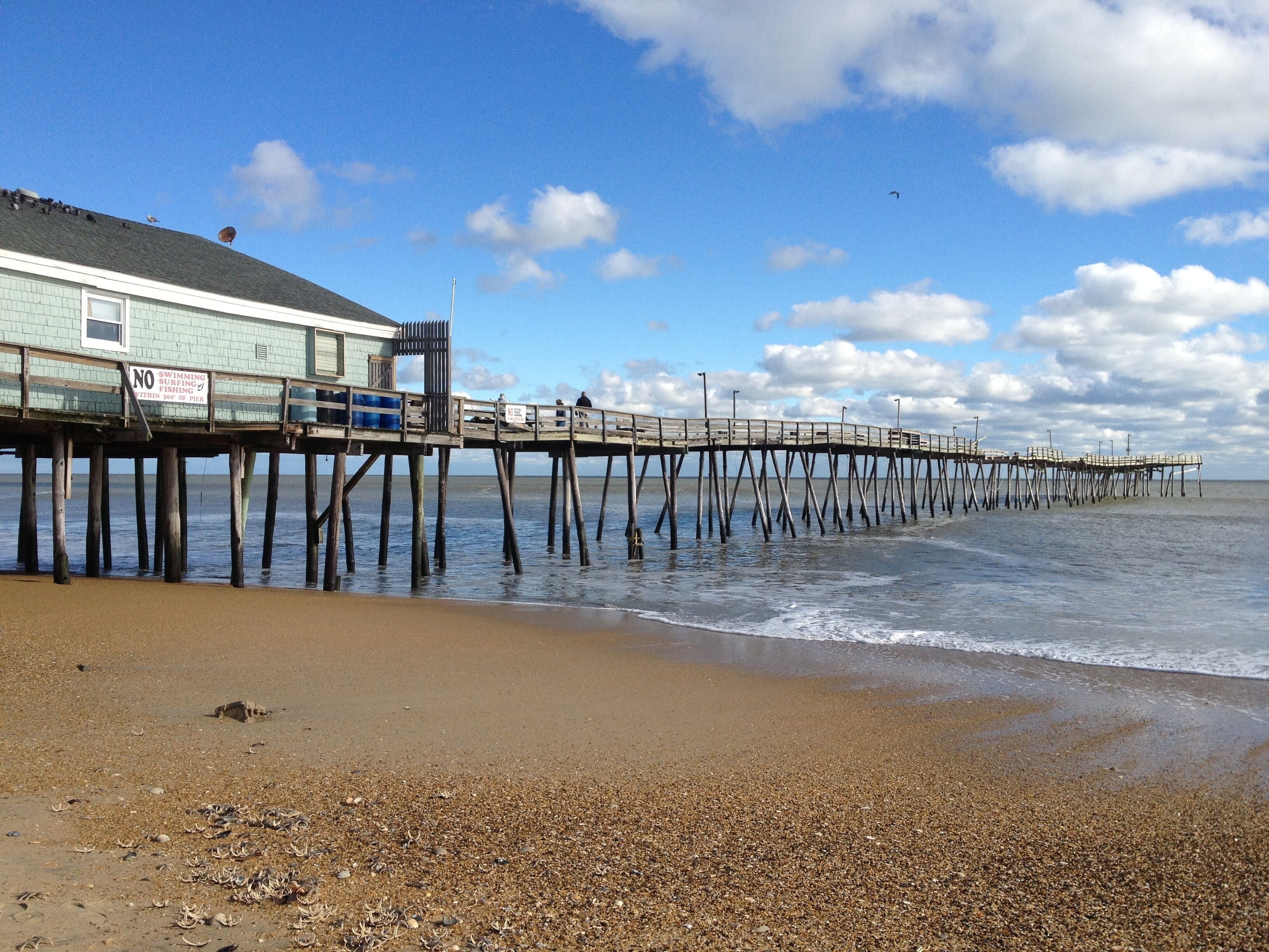 A beach with a wooden pier extending into the water, a building with a sign, and a clear sky.