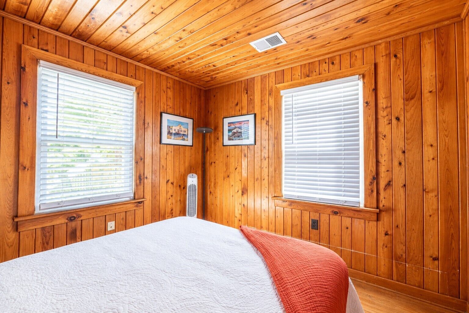 Une pièce aux murs recouverts de lambris en bois, un lit avec une couverture blanche et rouge, et deux fenêtres équipées de stores.