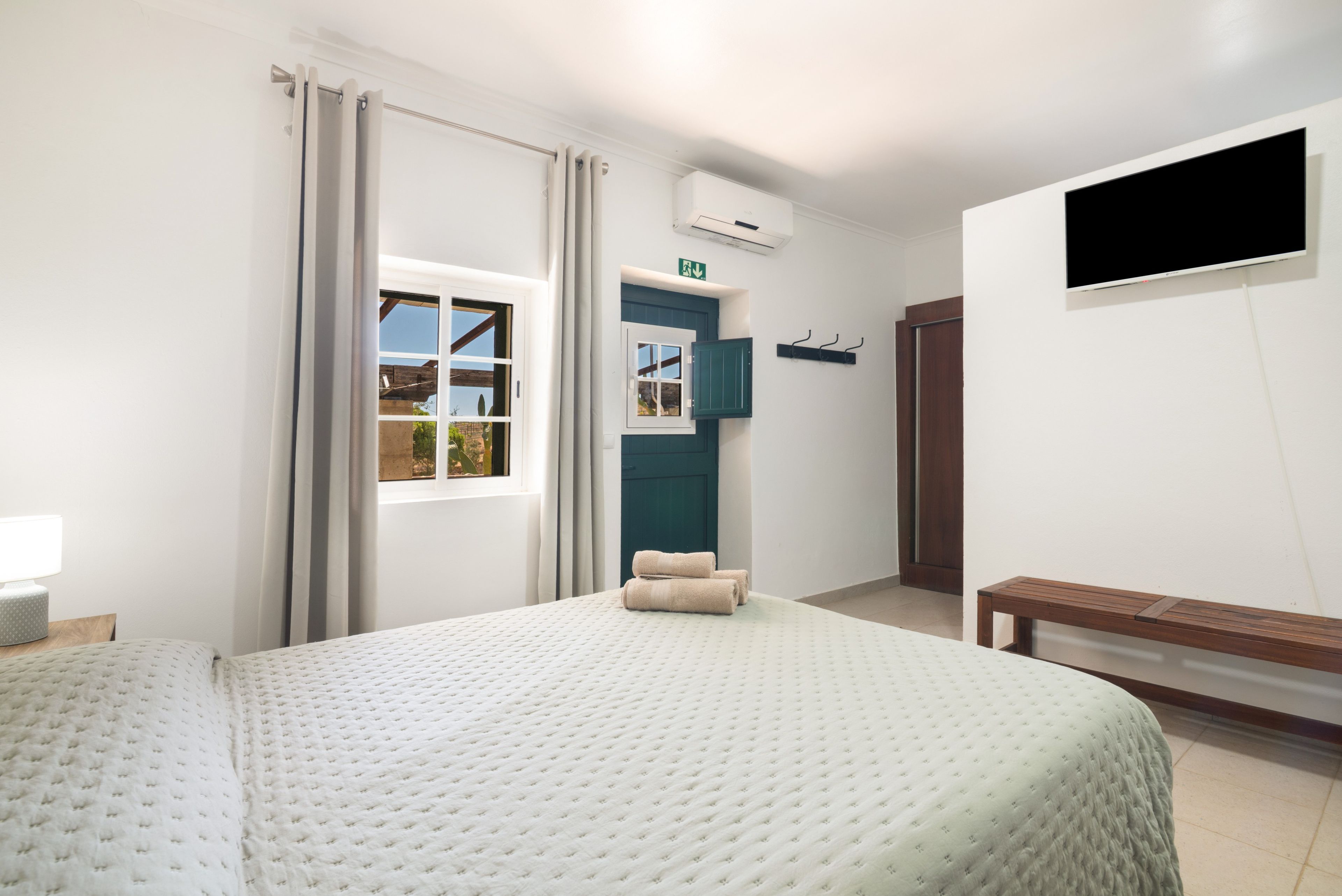 A hotel room with a large bed, a window with curtains, a TV mounted on the wall, and a bench.