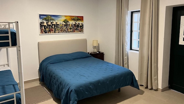 Une chambre avec un lit superposé, un lit simple avec une couvre-lit bleue, une table de chevet avec une lampe, une fenêtre avec des rideaux et un tableau accroché au mur.