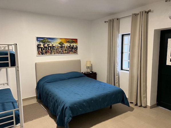 Een slaapkamer met een stapelbed, een enkel bed met een blauwe spreidek, een nachtkastje met een lamp, een raam met gordijnen, en een schilderij aan de muur.
