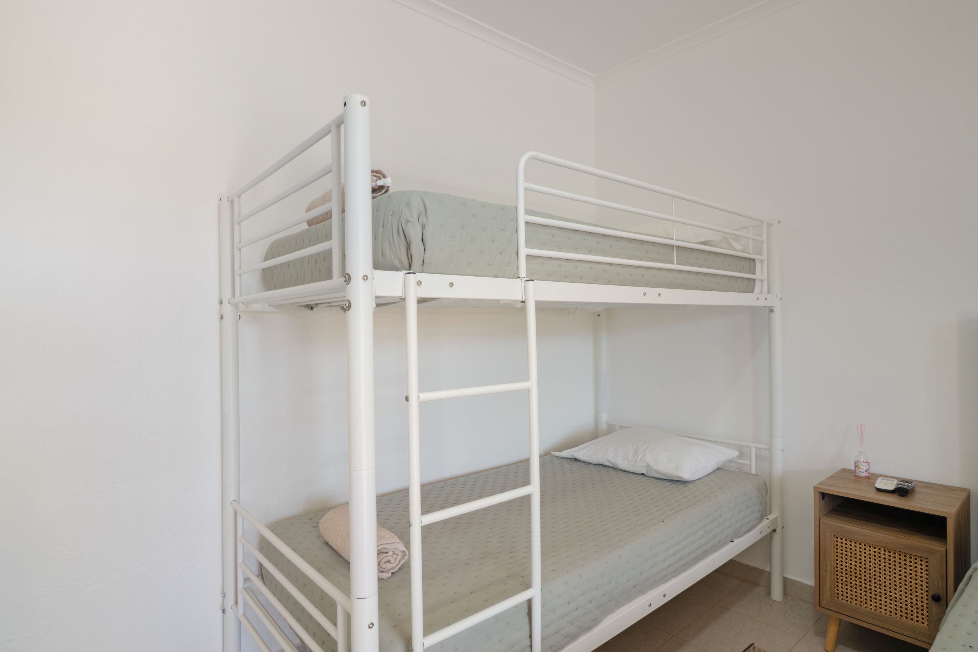 A bunk bed with a lower bunk and a single bed, a small wooden nightstand with a lamp, and a wall-mounted shelf.