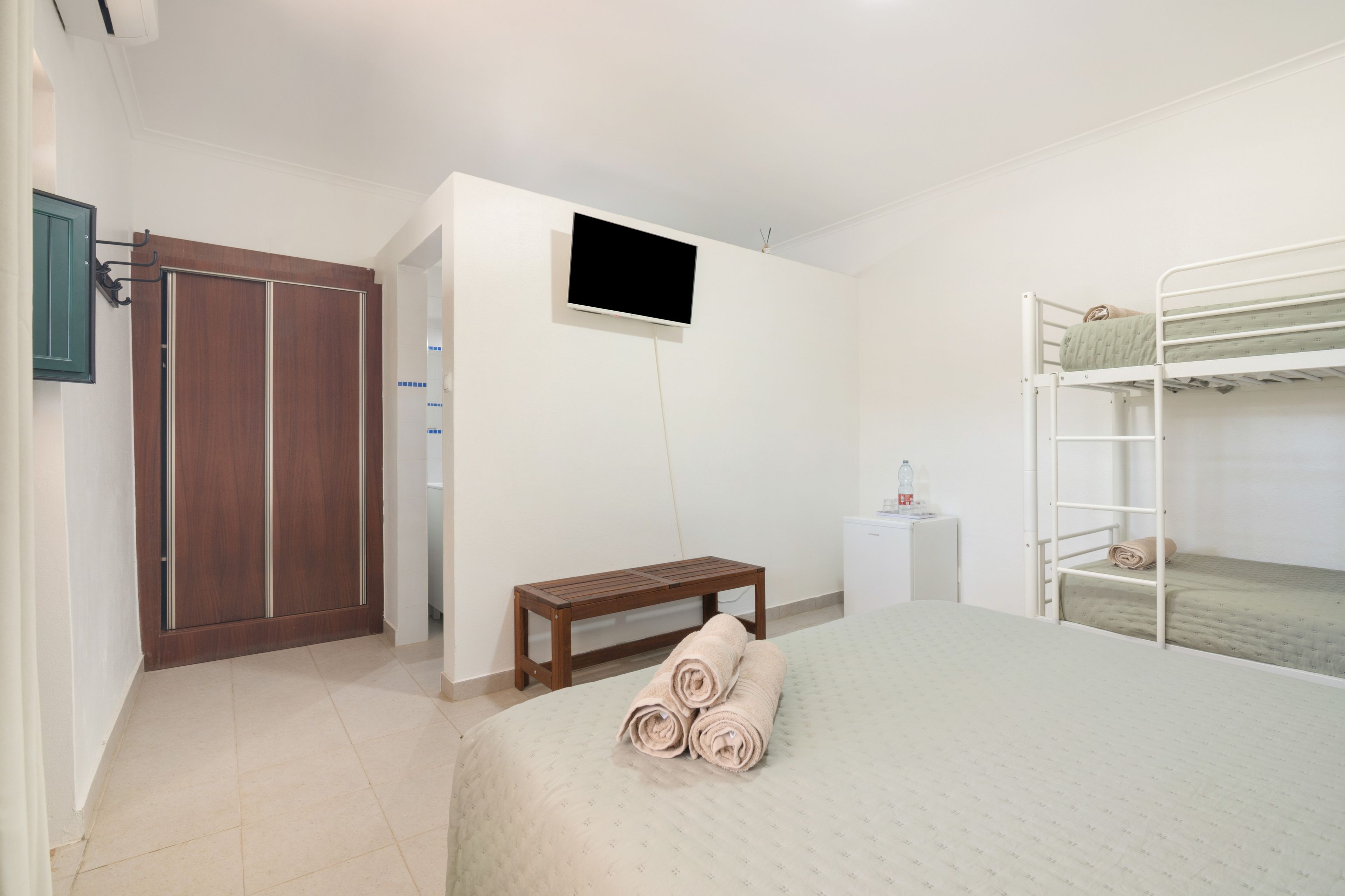 Een slaapkamer met een bed, een televisie, een kledingkast, een bankje en een stapelbed.