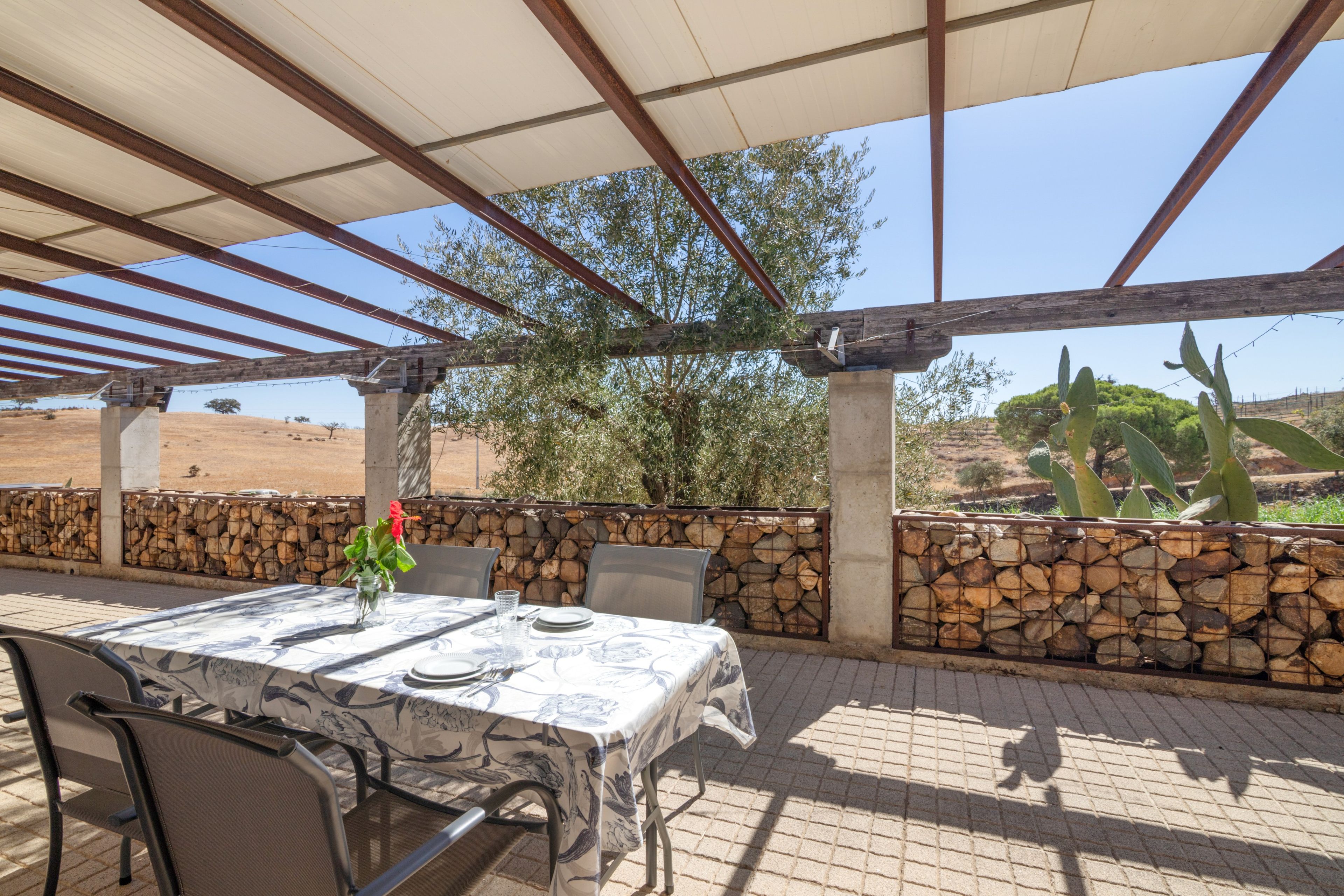 Une terrasse extérieure avec une table dressée pour le repas, entourée de murs en pierre et d’une pergola en bois.