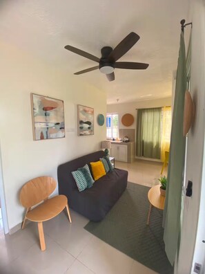 Ein Wohnzimmer mit einem dunklen Sofa, zwei Holzstühlen, einem Deckenventilator und abstrakter Wandkunst.