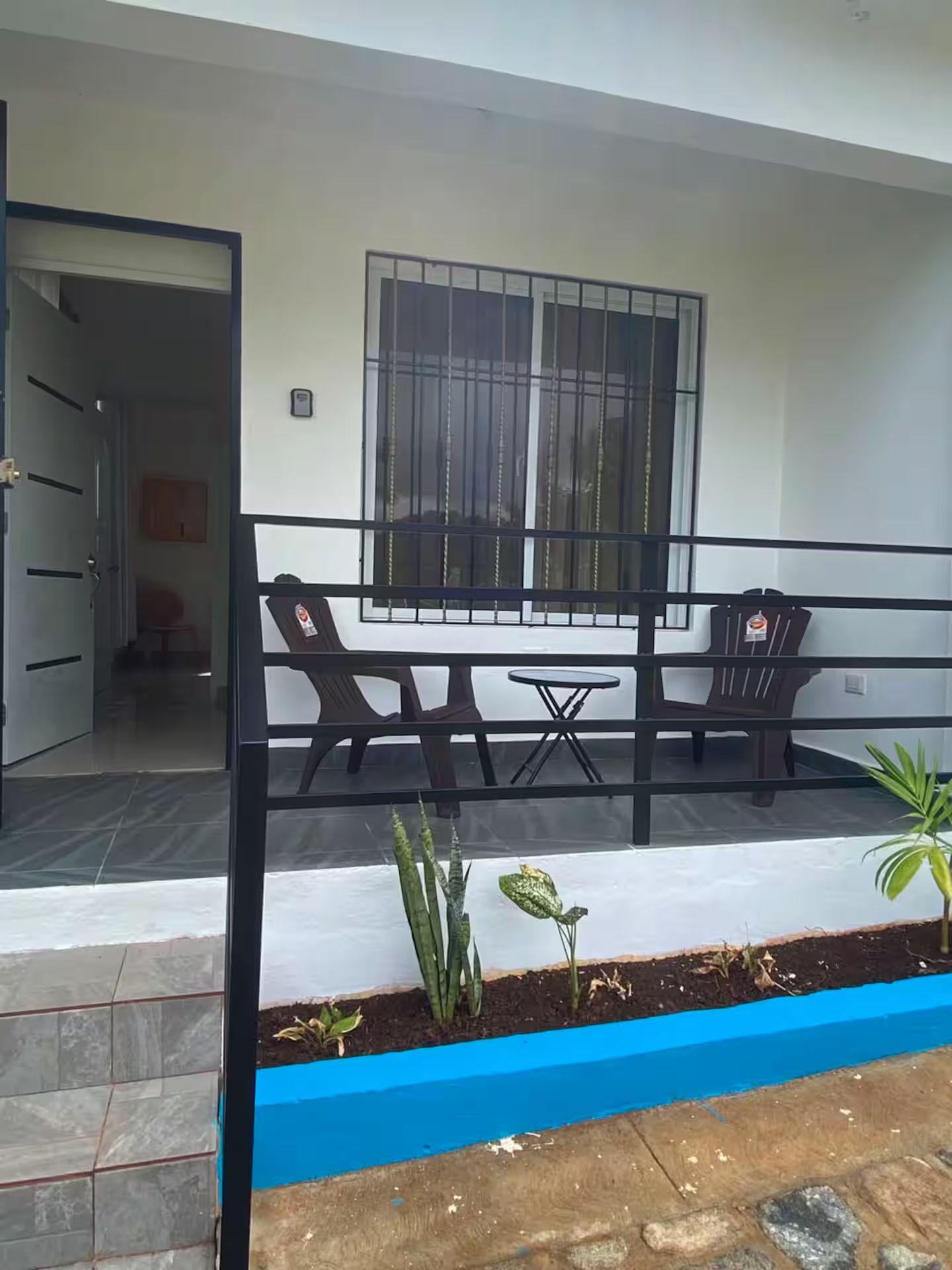 Un balcon avec une table et des chaises, une fenêtre avec des barreaux et un petit espace jardin avec des plantes.