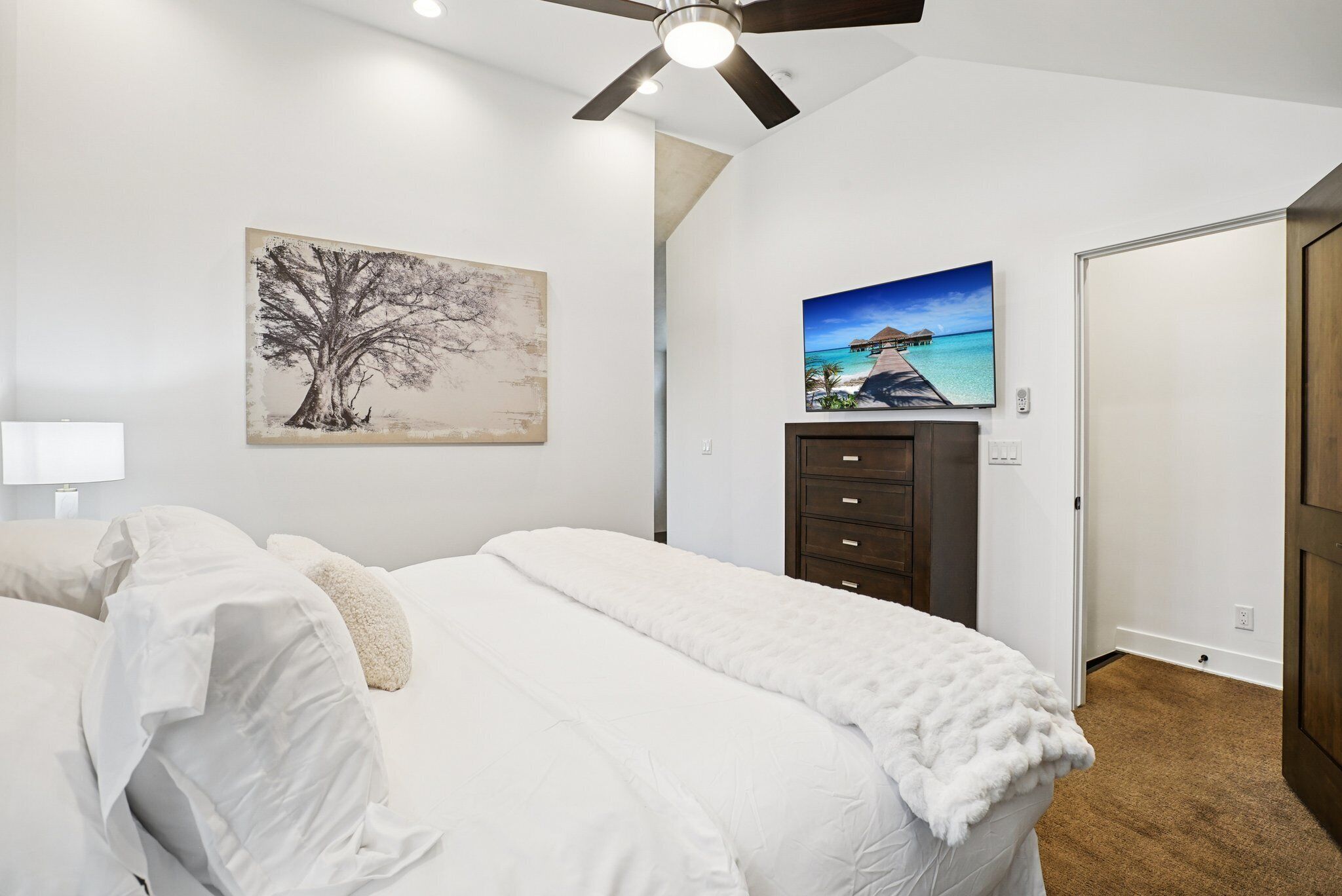 Un dormitorio con una cama, un ventilador de techo, una mesita de noche, una lámpara, un aparador y dos cuadros colgados en la pared.