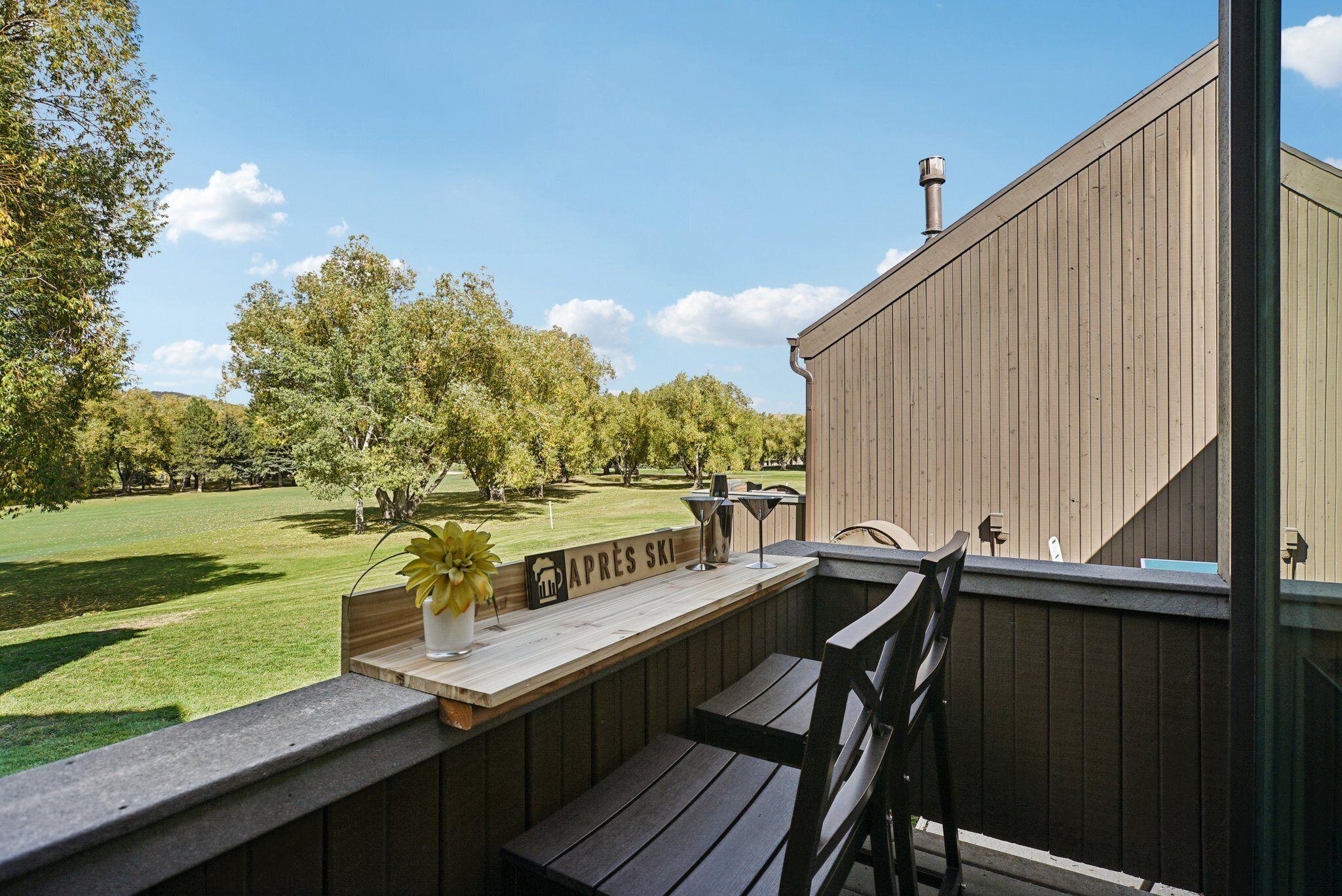 A wooden deck with a bench and a table, overlooking a grassy area with trees and a sign that reads "APRES SKI".
