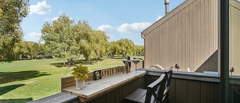 A wooden deck with a bench and a table, overlooking a grassy area with trees and a sign that reads "APRES SKI".