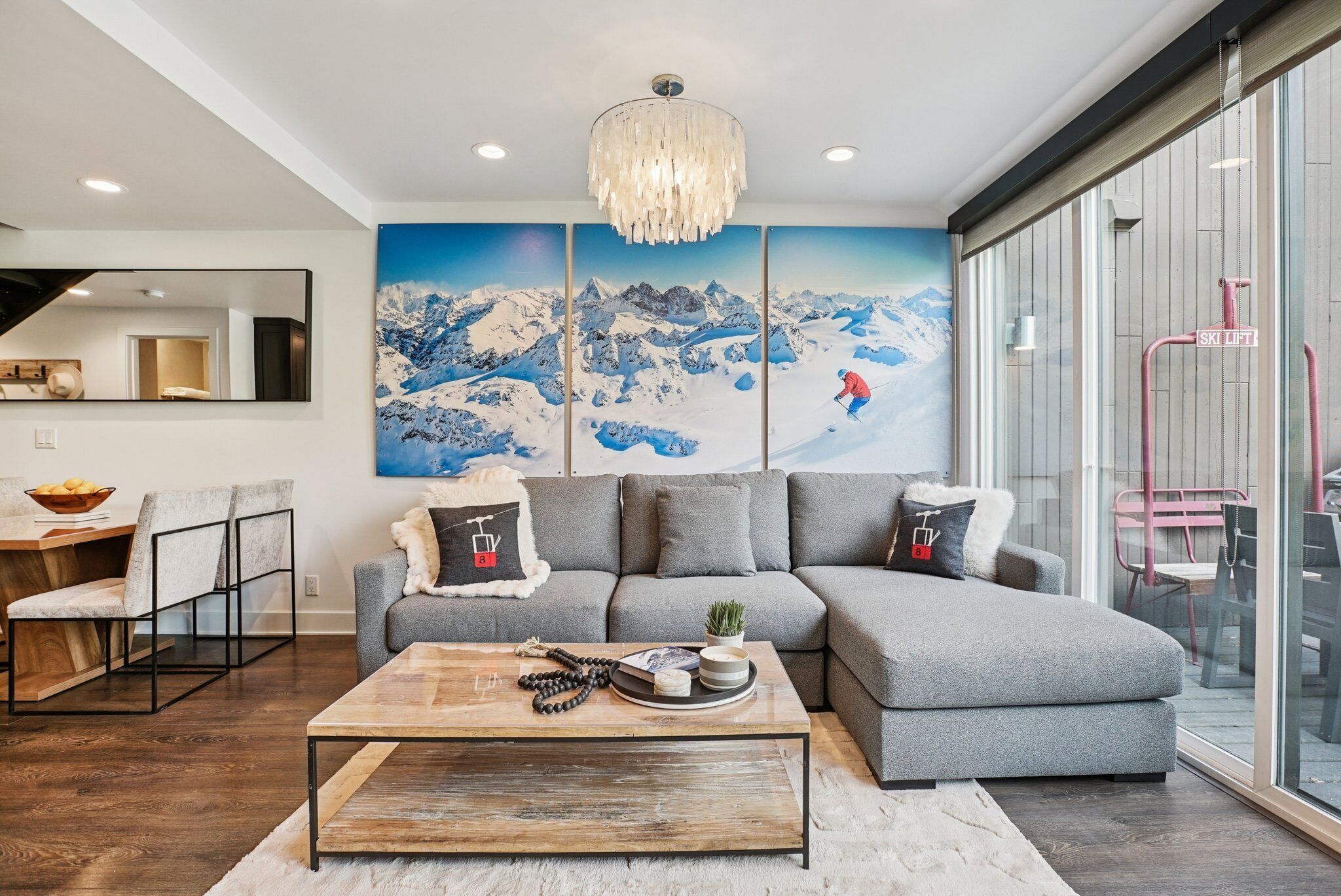 Un salon moderne avec un canapé d’angle gris, une table basse en bois et une grande fresque murale représentant un paysage de montagne enneigé.