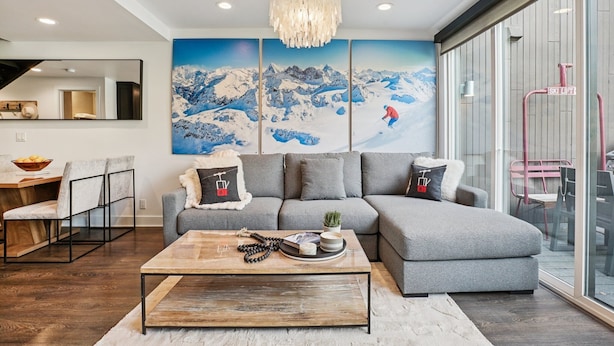 Una sala moderna con un sofá seccional gris, una mesa de centro de madera y un gran mural en la pared con una escena de montaña nevada.