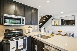 Una cocina moderna con armarios oscuros, electrodomésticos de acero inoxidable, un lavamanos doble y una escalera que sube a un nivel superior.