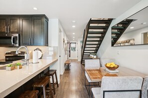 A modern kitchen with dark cabinets, a stainless steel microwave, and a sink. There is a wooden dining table with chairs and a bowl of lemons. A staircase with wooden steps leads to another level. - Three Kings Luxury Townhome 2BR Walk to Ski - Spa Inspired Bath (Park City)
