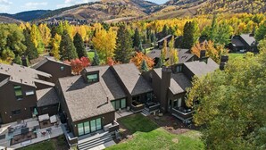 Un área residencial moderna con varias casas con tejados inclinados y grandes ventanales, rodeada de árboles de colores otoñales y un terreno montañoso al fondo.