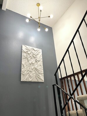 Une pièce avec un mur gris à texture, une œuvre d’art abstraite blanche, une rampe d’escalier en métal noir et un luminaire au plafond.