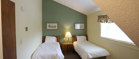 A small, single-room hotel room with two beds, a nightstand with a lamp, a window with a view, and a door.