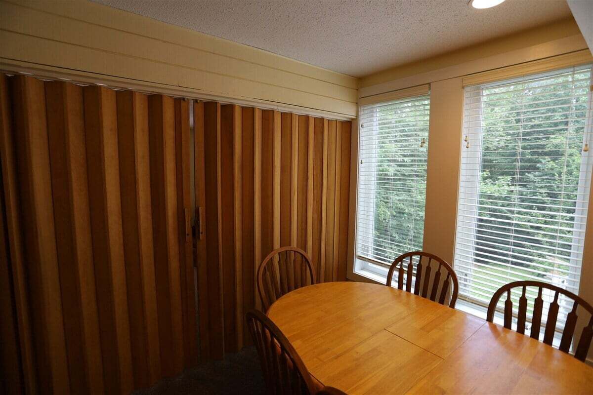 Ein Essbereich mit Holztisch und Stühlen, großen Fenstern mit Jalousien und einer Deckenleuchte.