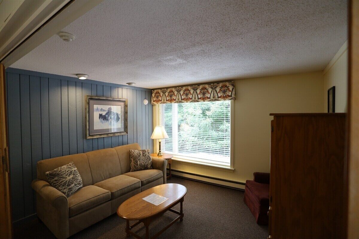 A living room with a sofa, a coffee table, a lamp, a window with blinds, and a framed picture on the wall.